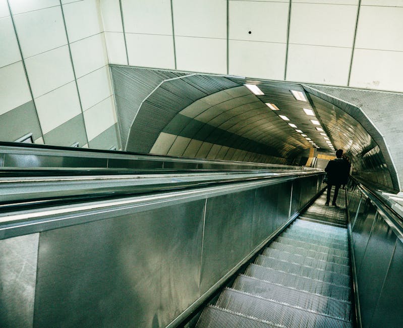 An underground escalator with metallic steps and side panels is descending into a tunnel. The tunnel's interior features smooth, curved metallic walls and ceiling with evenly spaced rectangular lights illuminating the space. To the right of the escalator, a person wearing dark clothing and a backpack is standing near the top, facing away from the camera. The escalator is situated inside a modern, tiled building with a minimalist design, and the surrounding area is clean and well-lit. This setting is relevant to house removals and furniture transport, illustrating the logistics involved in moving items through underground routes such as the Rotherhithe Tunnel, which [COMPANY_NAME], experts in removals, may access for efficient home relocation services.
