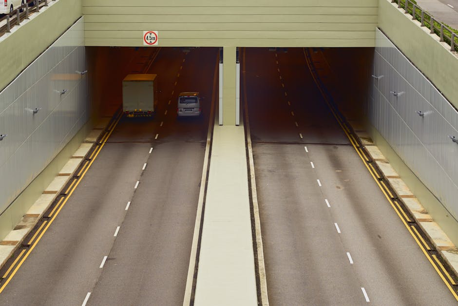 A view of a dual carriageway passing underneath a pedestrian bridge in a UK urban area, with clear lane markings and a 4.5-meter height restriction sign posted overhead. On the left lane, a small silver van is approaching the tunnel entrance, while a larger truck is seen further ahead. The right lane appears empty. The roadway is flanked by grey metal panels and green railings, with a pavement running parallel on both sides. This scene depicts a typical route used during house removals and furniture transport in the Rotherhithe area, relevant to loading, packing, and moving logistics handled by Man with Van Rotherhithe, who provides professional removals services in SE16. The environment is well-lit with natural daylight, emphasizing the urban infrastructure necessary for efficient home relocation.