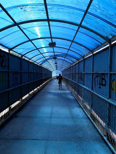 An underground escalator with metallic steps and side panels is descending into a tunnel. The tunnel's interior features smooth, curved metallic walls and ceiling with evenly spaced rectangular lights illuminating the space. To the right of the escalator, a person wearing dark clothing and a backpack is standing near the top, facing away from the camera. The escalator is situated inside a modern, tiled building with a minimalist design, and the surrounding area is clean and well-lit. This setting is relevant to house removals and furniture transport, illustrating the logistics involved in moving items through underground routes such as the Rotherhithe Tunnel, which [COMPANY_NAME], experts in removals, may access for efficient home relocation services.
