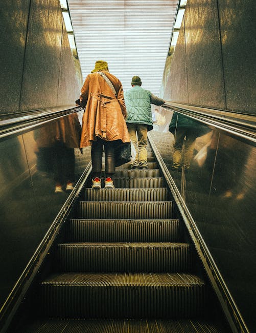 Inside a modern building with a metallic and glass structure, two individuals are seen ascending an escalator, viewed from the bottom. One person is wearing a beige coat, black pants, and red sneakers, while the other wears a green jacket with a hood, beige trousers, and brown shoes. The escalator is located adjacent to a polished black wall on the left side, and the ceiling above is illuminated with bright lighting, creating a well-lit environment. The surrounding environment suggests an urban setting, possibly a train station or commercial building, associated with house removals and relocation logistics. This image depicts the process of moving or travelling within a city, supporting professional moving services such as those offered by Man with Van Rotherhithe, especially in contexts involving stairs or lifts for home or furniture transport.
