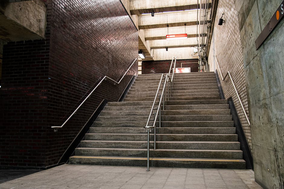 A concrete staircase with metal handrails ascending inside a building, likely part of a residential or commercial property in Rotherhithe. The staircase has multiple steps made of concrete, with some carrying cardboard boxes wrapped in plastic, possibly for a home relocation or furniture transport process. The surrounding environment includes a dark brick wall on one side, a concrete wall on the other, and an overhead ceiling with visible pipes and lighting fixtures. At the top of the stairs, a hallway or landing area is visible, with additional doors or entrances. The scene suggests an indoor loading or moving situation, with a focus on careful handling and logistical organization typical of professional removals carried out by [COMPANY_NAME], supporting services like packing and stairs or lift access for efficient residential or office moves.