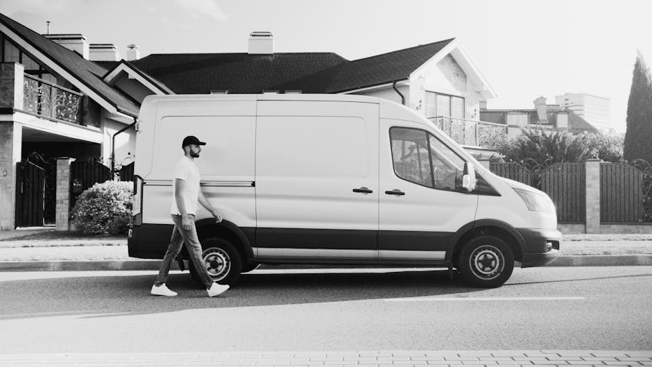 A person wearing a dark jumpsuit and red shoes stands outside in daylight, loading a large green box into the open rear cargo area of a white van parked on a paved street. The van's back doors are wid
