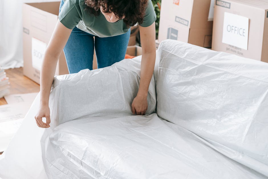 A person in casual clothing is seen wrapping a large, upholstered sofa with white packing material, with one end already covered and the other being carefully prepared for protection during removals. The sofa is positioned inside a room, likely a living area, with several cardboard boxes and packing supplies, including plastic wrapping and packing paper, visible in the background. The individual is leaning over and using their hands to secure the wrapping around the furniture, ensuring it remains protected for safe transportation. The scene captures the packing and moving process essential to house removals, with a focus on safeguarding furniture during a home relocation. Additionally, a moving van or storage area might be nearby, though not visible in this particular shot. The setup appears well-organized, with adequate padding and packing materials to minimize damage during the furniture transport handled by Man with Van Rotherhithe.