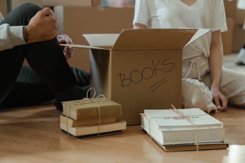 A person sitting on a wooden floor beside a cardboard box labeled 'BOOKS' with black marker, surrounded by neatly stacked books tied with twine and additional books nearby, with another individual sitting nearby wearing light-colored clothing, in a home environment with natural lighting. The scene depicts the packing process involved in a house relocation or moving service, with the box and books positioned close to the open doorway and adjacent to a moving truck or van outside, supporting the context of furniture transport and packing for home removals managed by Man with Van Rotherhithe.