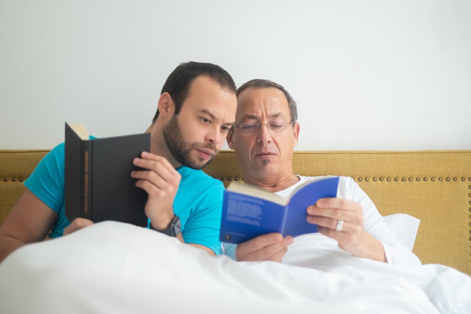 Two men sitting on a bed inside a room, with a mustard-colored upholstered headboard behind them. The man on the left, wearing a bright blue t-shirt, is holding a small black book with a beige spine and appears to be looking at it with a focused expression. The man on the right, dressed in a white shirt, is holding an open blue book and reading it with a slightly puzzled look. The bed has white bedding, and there is a plain white wall in the background. The scene suggests a casual moment of reading or studying indoors. This image may be associated with house removals and moving services, such as packing or preparing for a home relocation, as provided by Man with Van Rotherhithe.