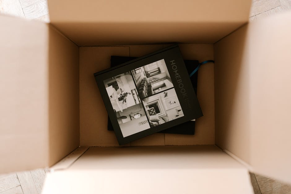 A person sitting on a wooden floor beside a cardboard box labeled 'BOOKS' with black marker, surrounded by neatly stacked books tied with twine and additional books nearby, with another individual sitting nearby wearing light-colored clothing, in a home environment with natural lighting. The scene depicts the packing process involved in a house relocation or moving service, with the box and books positioned close to the open doorway and adjacent to a moving truck or van outside, supporting the context of furniture transport and packing for home removals managed by Man with Van Rotherhithe.