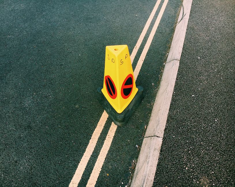 A yellow traffic cone with black rubber base placed on a double yellow line on a tarmac road, near a curb made of concrete. The cone has some handwritten markings in black ink on its surface. The road surface is dark and slightly textured, with the double yellow lines indicating a no-parking zone. In the background, part of the curb and the edge of the pavement are visible. This scene is representative of parking restrictions that may be relevant during house removals or furniture transport, highlighting the importance of proper planning and adherence to local parking regulations to avoid fines during moving day. The presence of the cone suggests a temporary restriction, often seen in urban environments like Rotherhithe, where moving companies such as Man with Van Rotherhithe operate within regulated areas to facilitate home relocation and logistics.