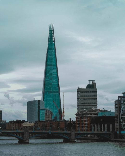 A tall, modern skyscraper with a glass exterior reflecting the cloudy sky dominates the city skyline, with several other high-rise buildings of varying architectural styles surrounding it. In the foreground, a bridge spans a river, with boats and water traffic visible below. The scene is set during daytime with overcast weather, and the buildings and river are part of the urban landscape associated with property and house removals in Southwark, Rotherhithe, as discussed on the Man with Van Rotherhithe website. The image highlights the city's contemporary development, relevant to relocation and furniture transport services in the area.
