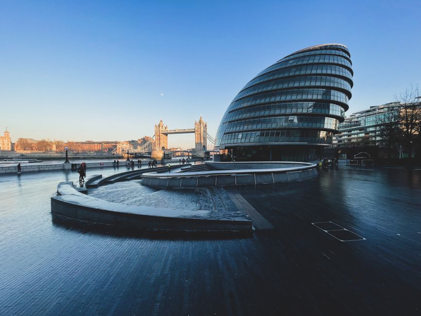 A modern, curved glass office building with multiple floors and horizontal windows, situated on a paved area beside a body of water in Rotherhithe. In the background, Tower Bridge with its iconic towers and suspension cables spans the River Thames, with a clear blue sky overhead. Several pedestrians are walking along the waterfront, some near the building and others closer to the bridge. The paved area features a circular water fountain or seating structure, with the ground appearing wet and reflecting the building and sky. The scene is well-lit by natural daylight, highlighting the architectural features and the surrounding environment. This location appears to be a popular area for public walking and sightseeing, close to key transport links and urban landmarks. Man with Van Rotherhithe may utilize this environment for local home relocations or furniture transport, with the prominent building and bridge serving as recognizable local references.