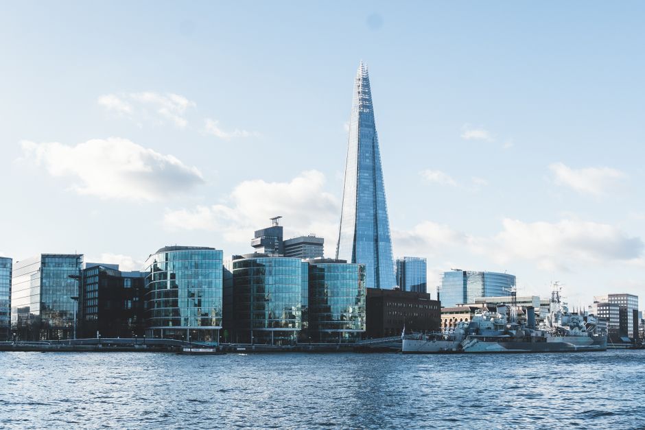 A tall, modern skyscraper with a glass exterior reflecting the cloudy sky dominates the city skyline, with several other high-rise buildings of varying architectural styles surrounding it. In the foreground, a bridge spans a river, with boats and water traffic visible below. The scene is set during daytime with overcast weather, and the buildings and river are part of the urban landscape associated with property and house removals in Southwark, Rotherhithe, as discussed on the Man with Van Rotherhithe website. The image highlights the city's contemporary development, relevant to relocation and furniture transport services in the area.