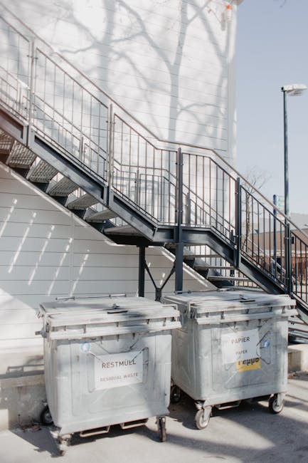 A row of large waste disposal bins and containers positioned outside a property, with four white recycling bins lined up next to a black waste container, all placed on a pavement area. The recycling b